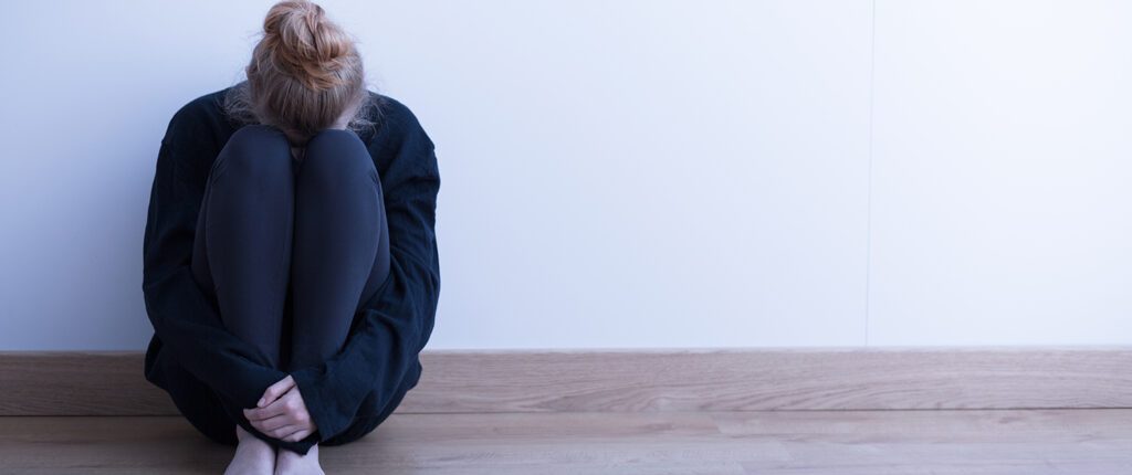 Zusammengekauerte Frau sitzt auf dem Boden an eine Wand gelehnt