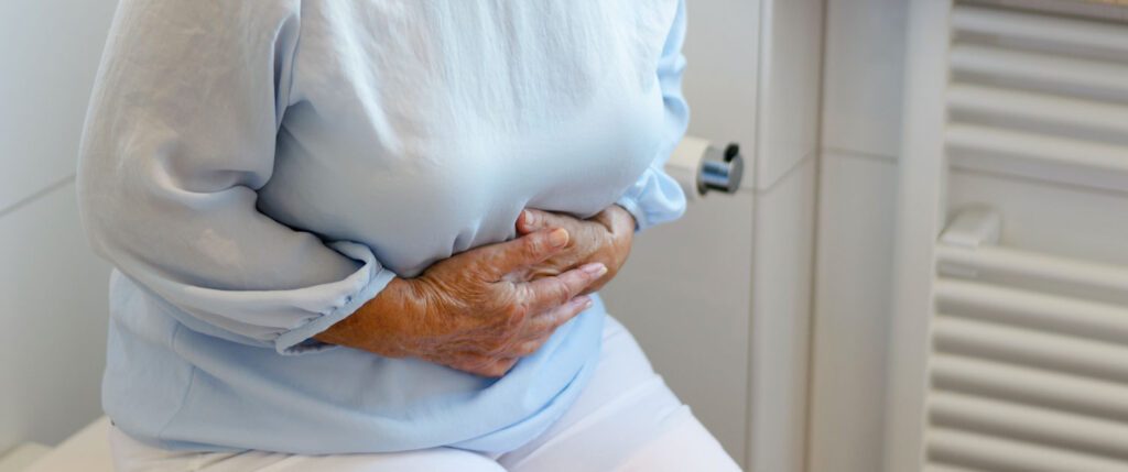 Eine auf dem geschlossenen Toilettendeckel sitzende Frau hält sich den Bauch.