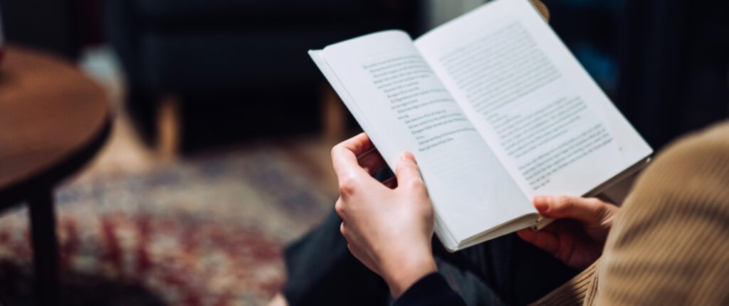 Cropped shot of young Asian woman taking a break from technology, relaxing and reading book on the sofa at a cozy home in the evening